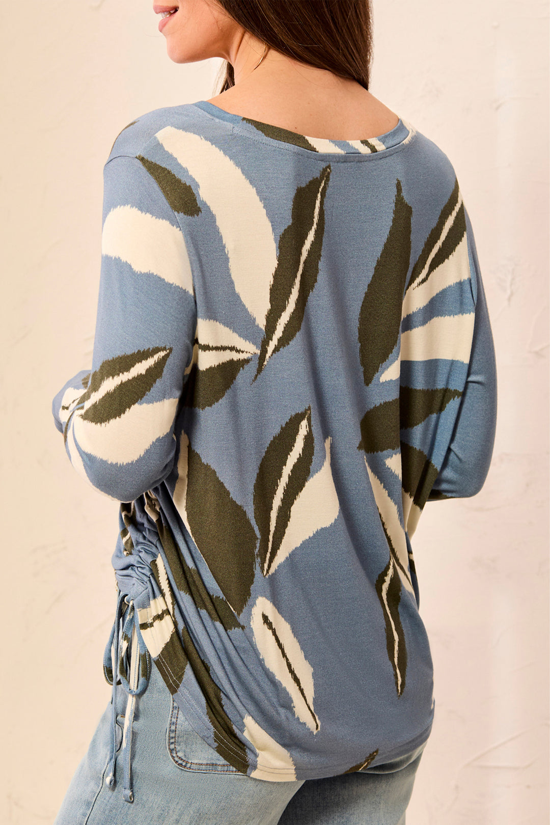 A woman wearing a blue knit top with a large leaf pattern in cream and olive green, shown from the back. The top has a round neckline, elbow-length sleeves, and a side tie detail at the back hem.