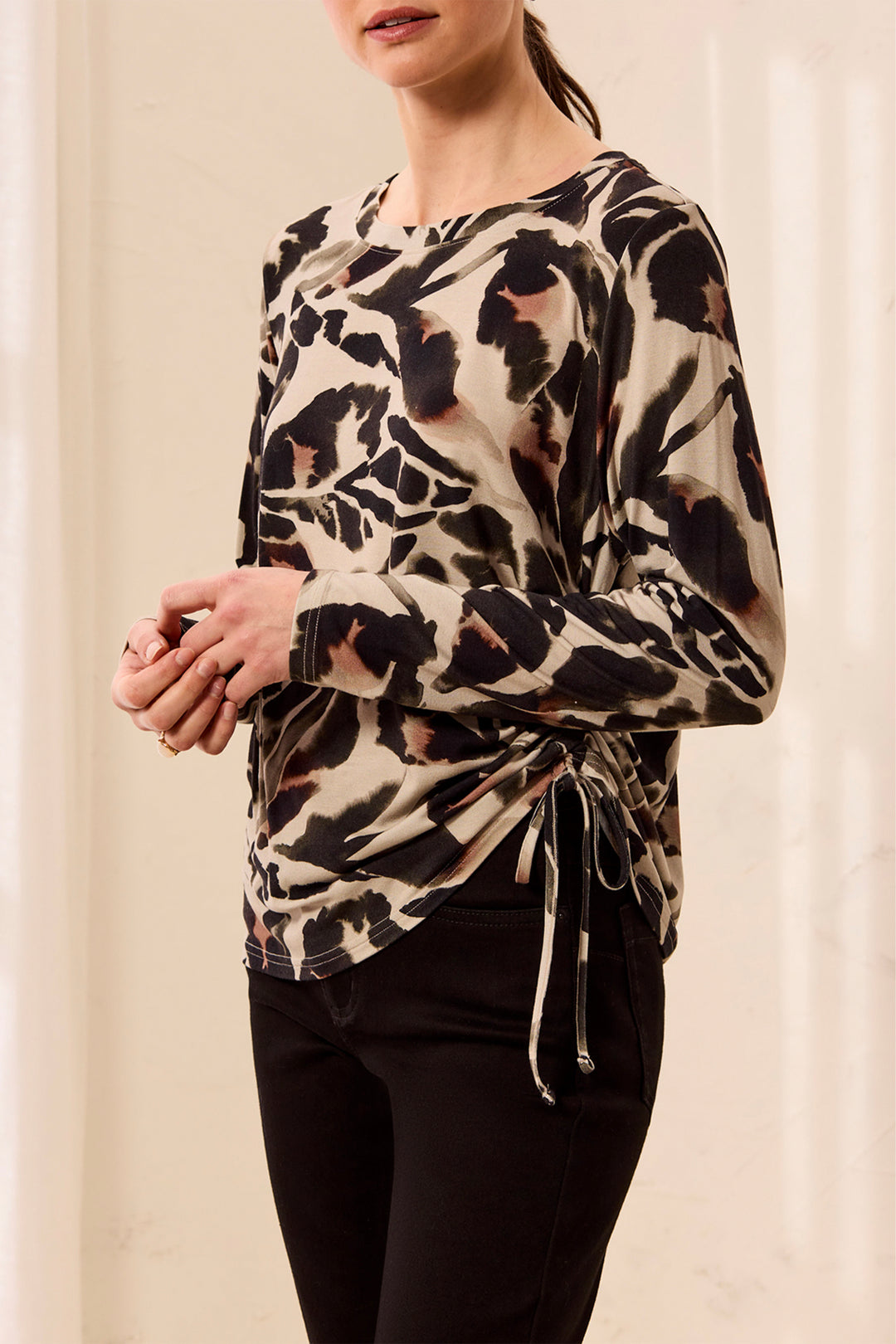 A woman wearing a long-sleeve beige blouse with a black and brown abstract animal print and a side tie detail, paired with black pants.