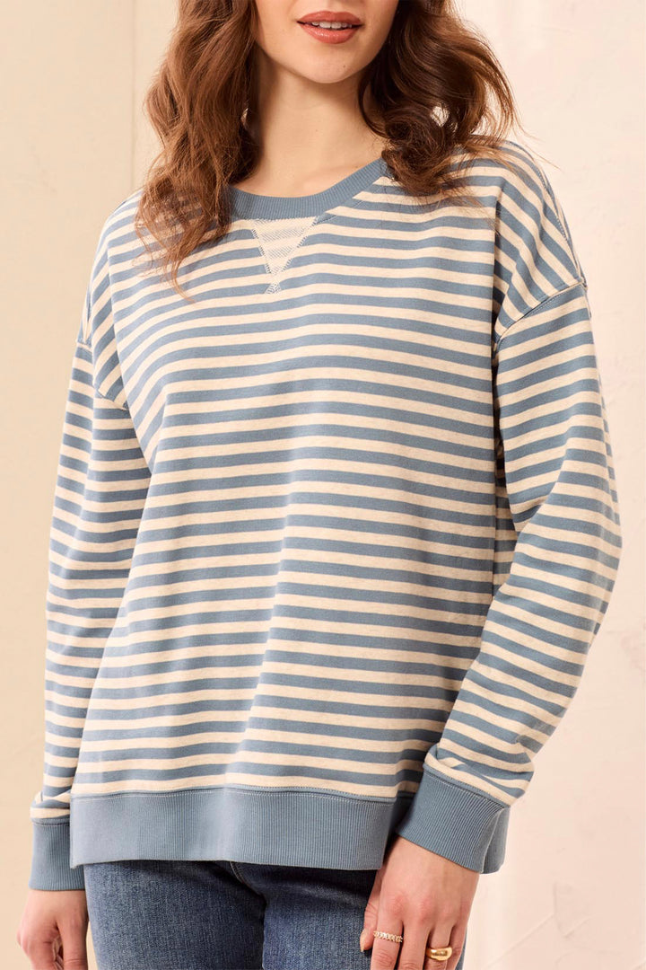 A woman wearing a long-sleeve crewneck sweatshirt with horizontal blue and cream stripes, paired with blue jeans, against a light beige background.