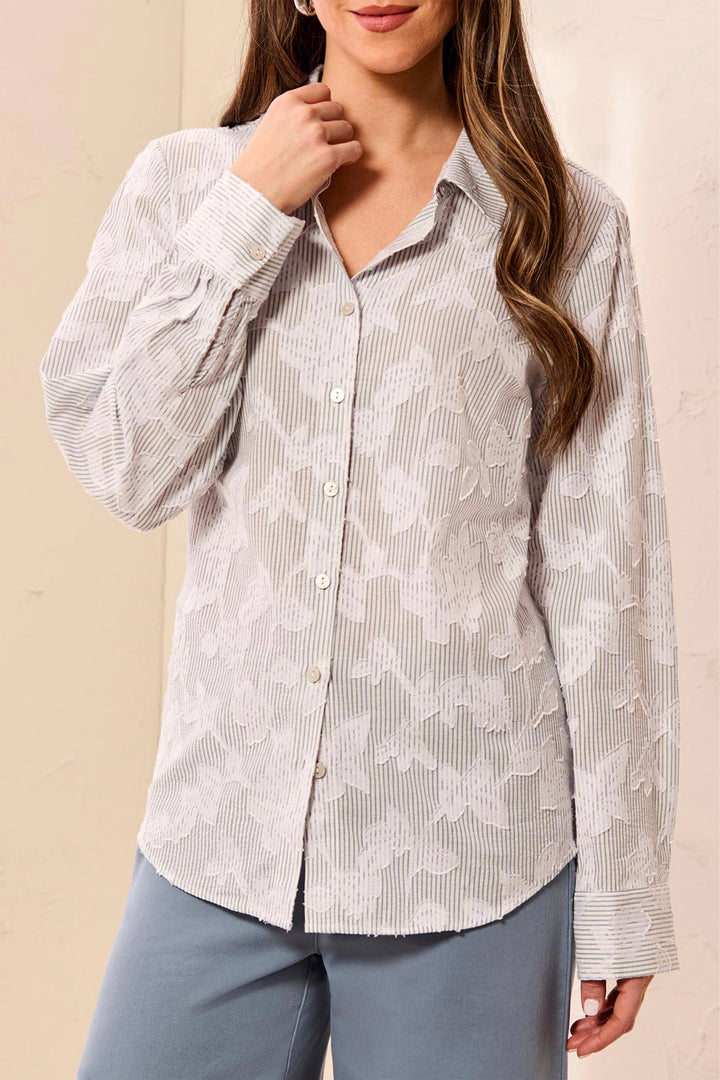 A woman wearing a light beige and white striped button-down blouse with a subtle floral jacquard pattern, paired with slate blue pants, set against a warm beige background.