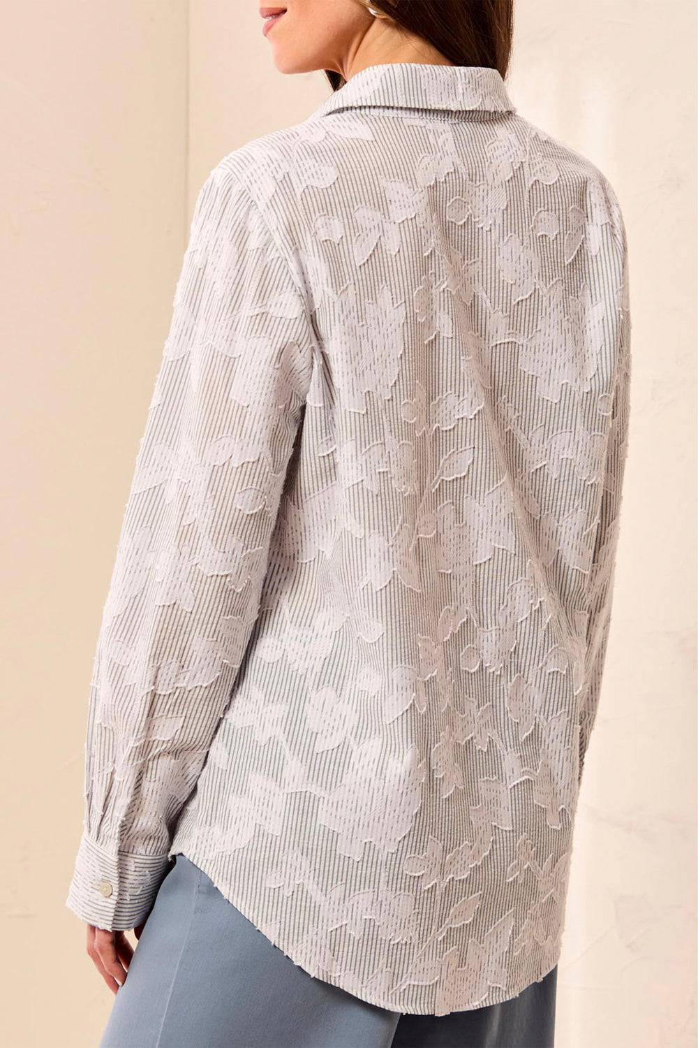 A woman shown from the back wearing a light gray and white striped button-up shirt with a raised floral lace-like overlay, paired with blue-gray pants.