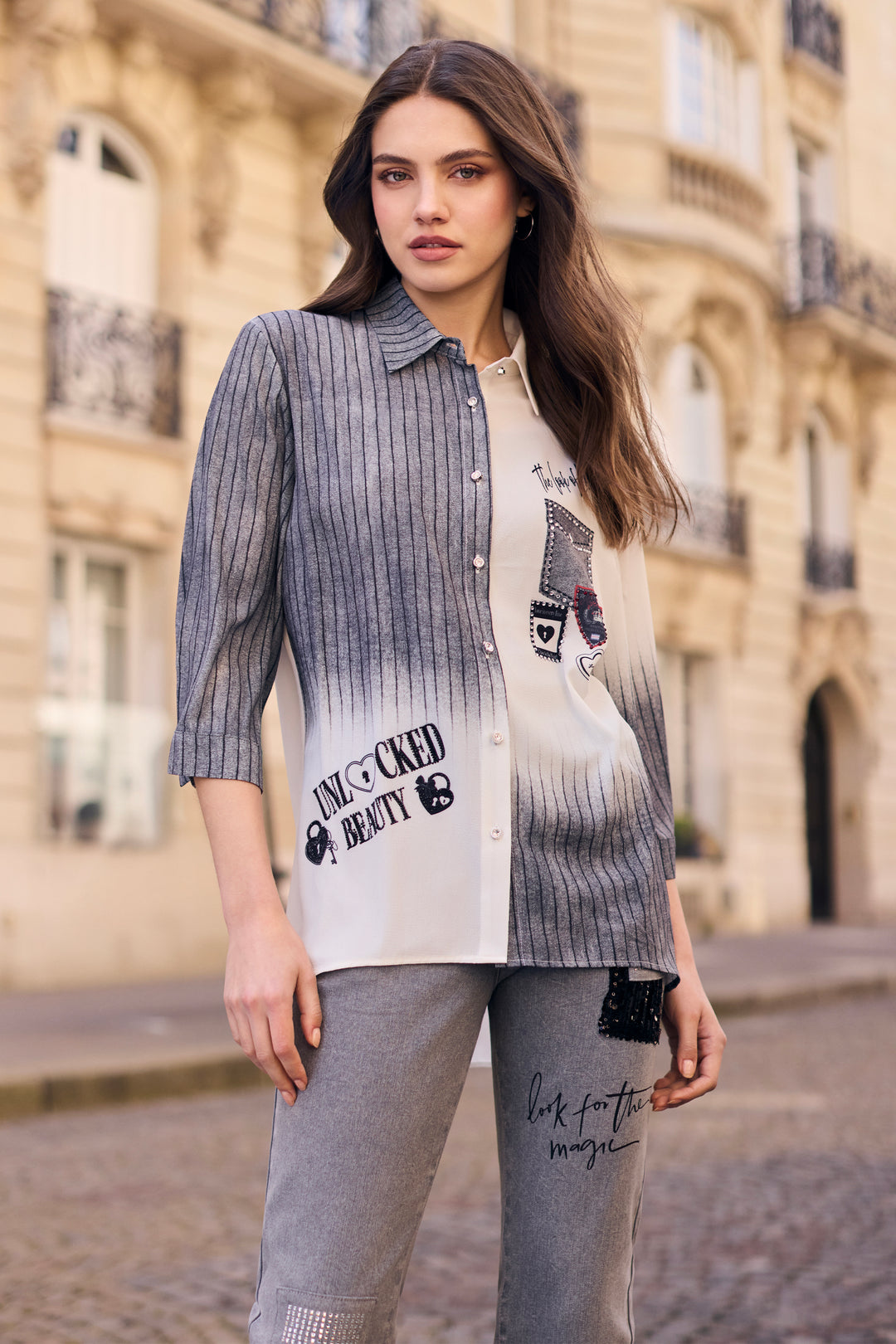 A woman stands outdoors in front of a beige stone building, wearing a long-sleeve button-front shirt with a gray-to-white ombré stripe pattern, black graphic text print, and gray jogger pants.