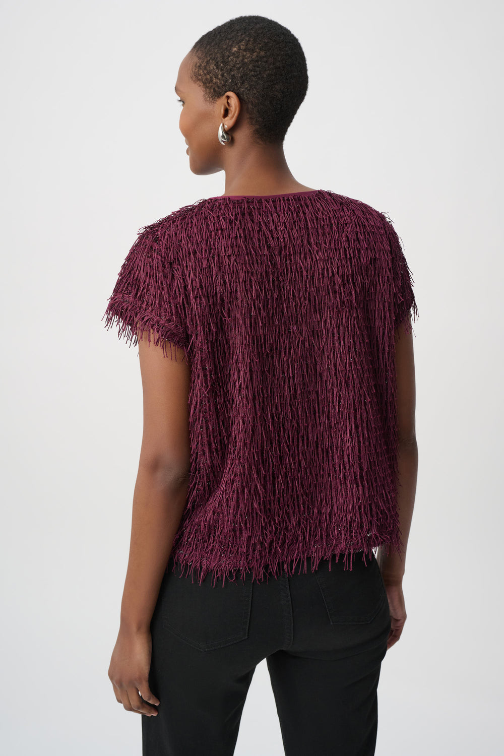 A woman shown from the back wearing a short-sleeved, fringed burgundy top and black pants against a plain light gray studio background.