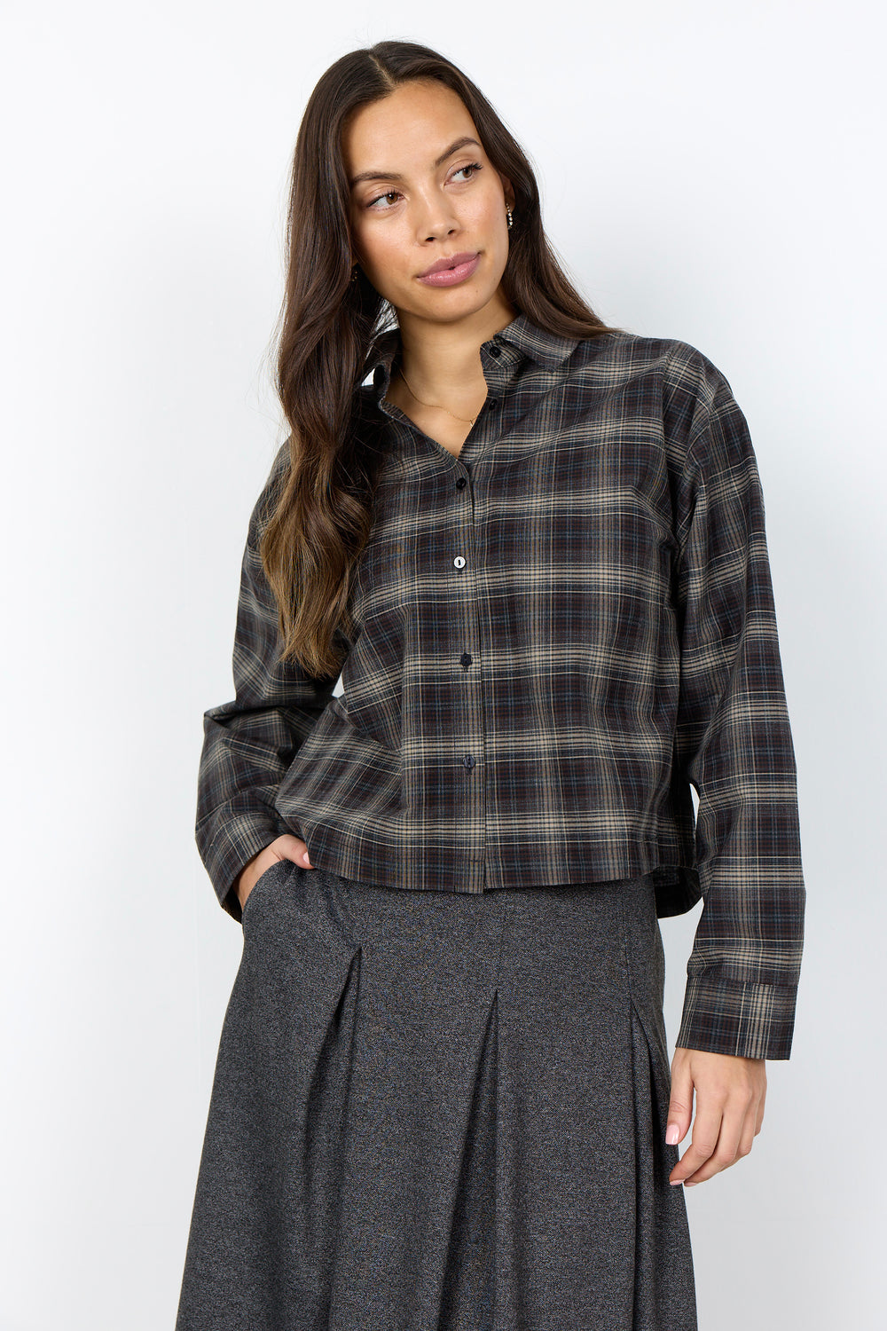 A woman modeling a dark brown and black plaid button-down shirt with a gray pleated skirt, standing against a plain white studio background.