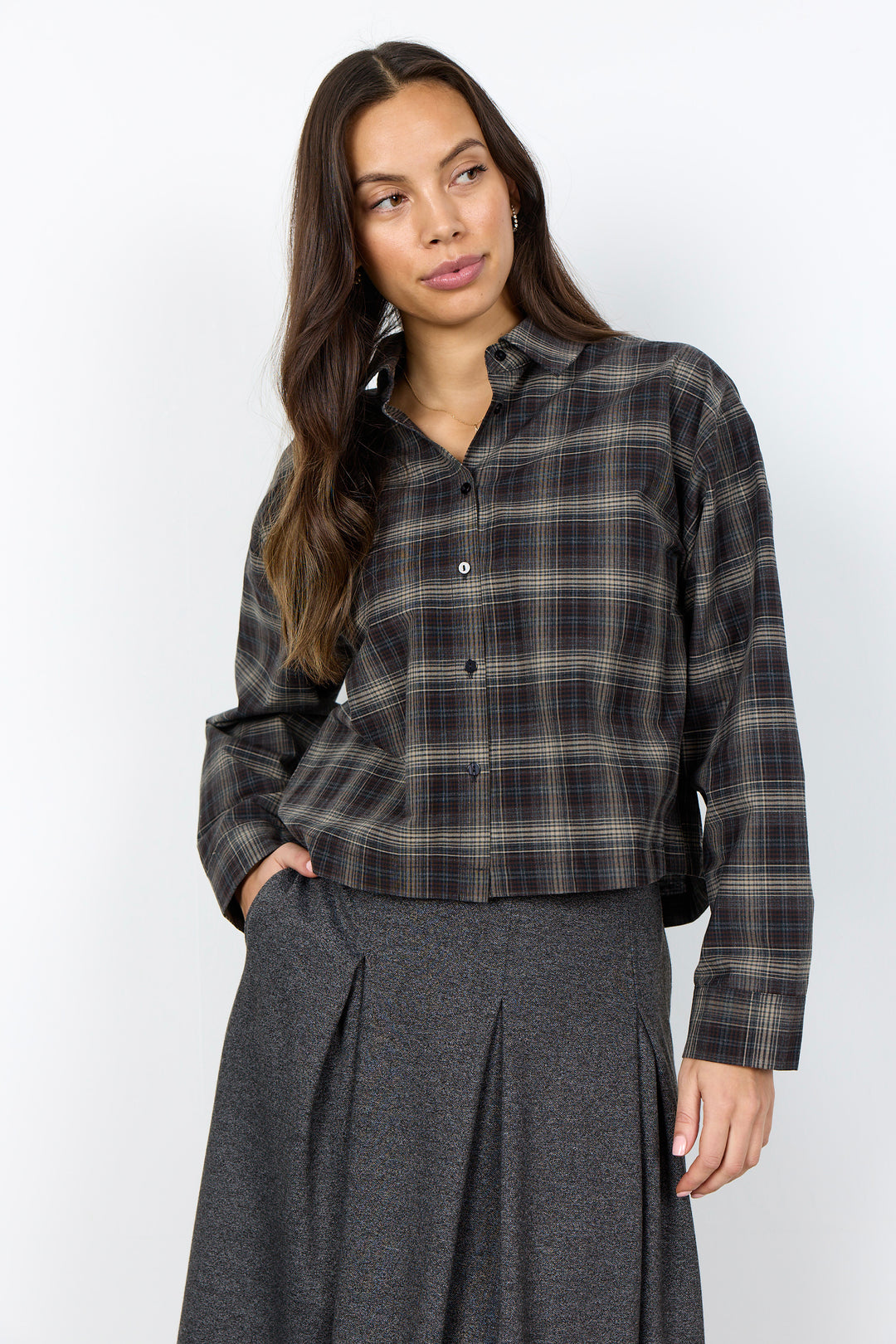 A woman modeling a dark brown and black plaid button-down shirt with a gray pleated skirt, standing against a plain white studio background.