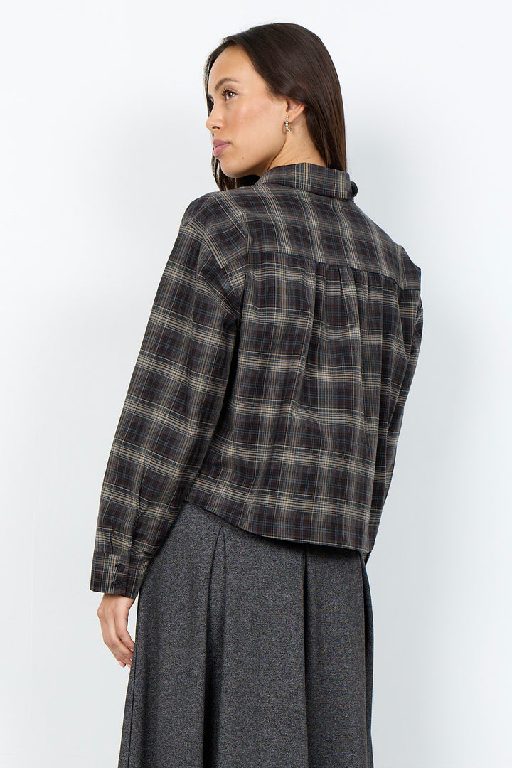 A woman shown from the back wearing a dark gray and black plaid button-up shirt and a dark gray skirt, photographed against a plain light gray studio background.