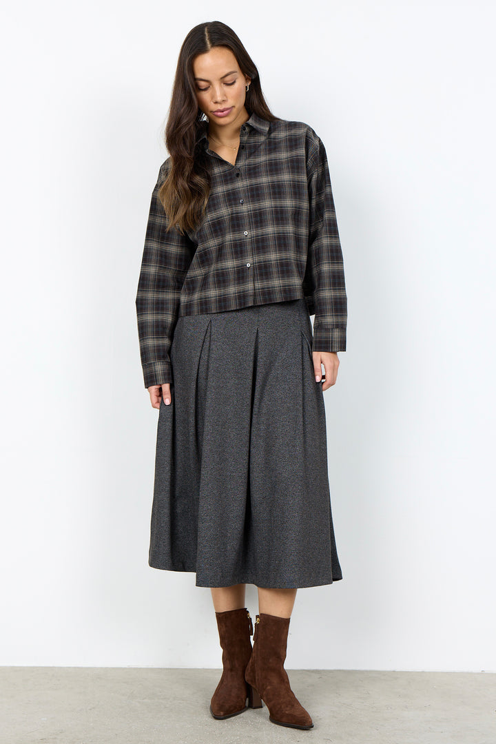 A woman stands against a plain light gray wall wearing a dark gray and black plaid button-up shirt, a dark gray pleated midi skirt, and brown ankle boots.