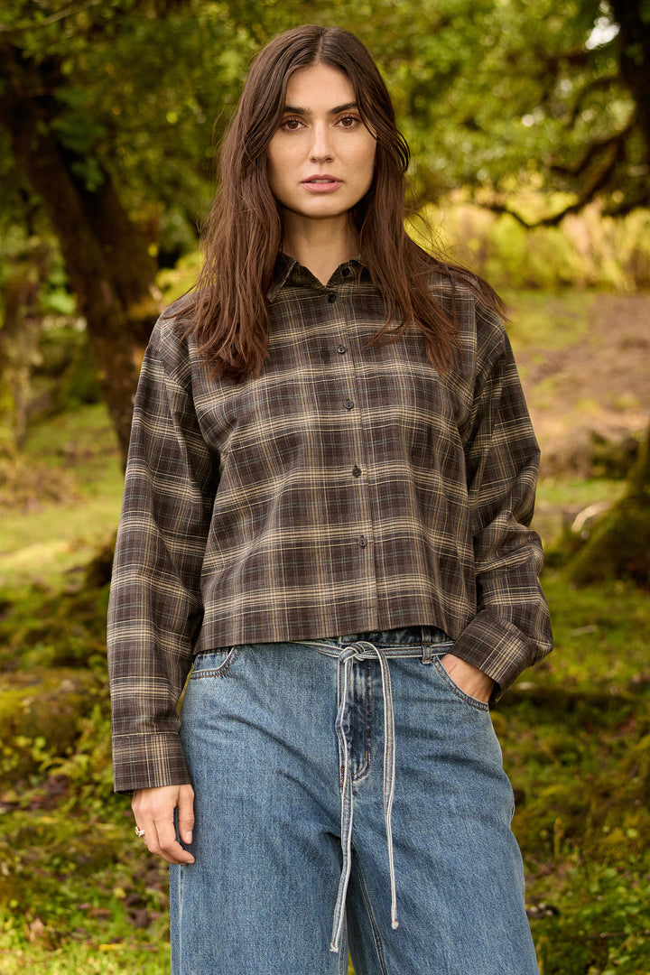 A woman standing outdoors in a wooded setting, wearing a cropped long-sleeve button-up plaid shirt in brown and beige tones with blue jeans.