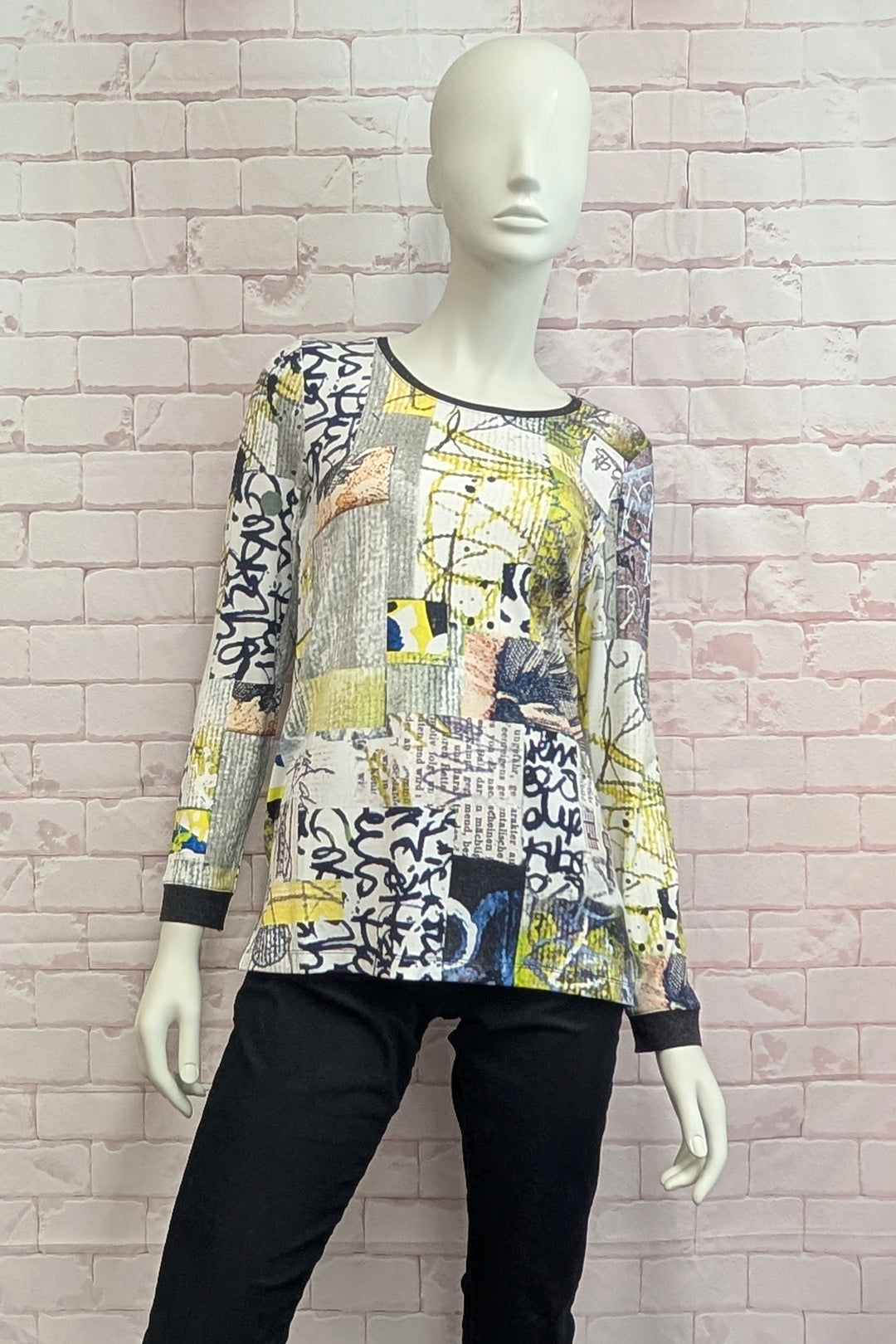 A long-sleeve patchwork top with an abstract collage print in yellow, black, white, and gray, displayed on a female mannequin against a pink brick wall background.
