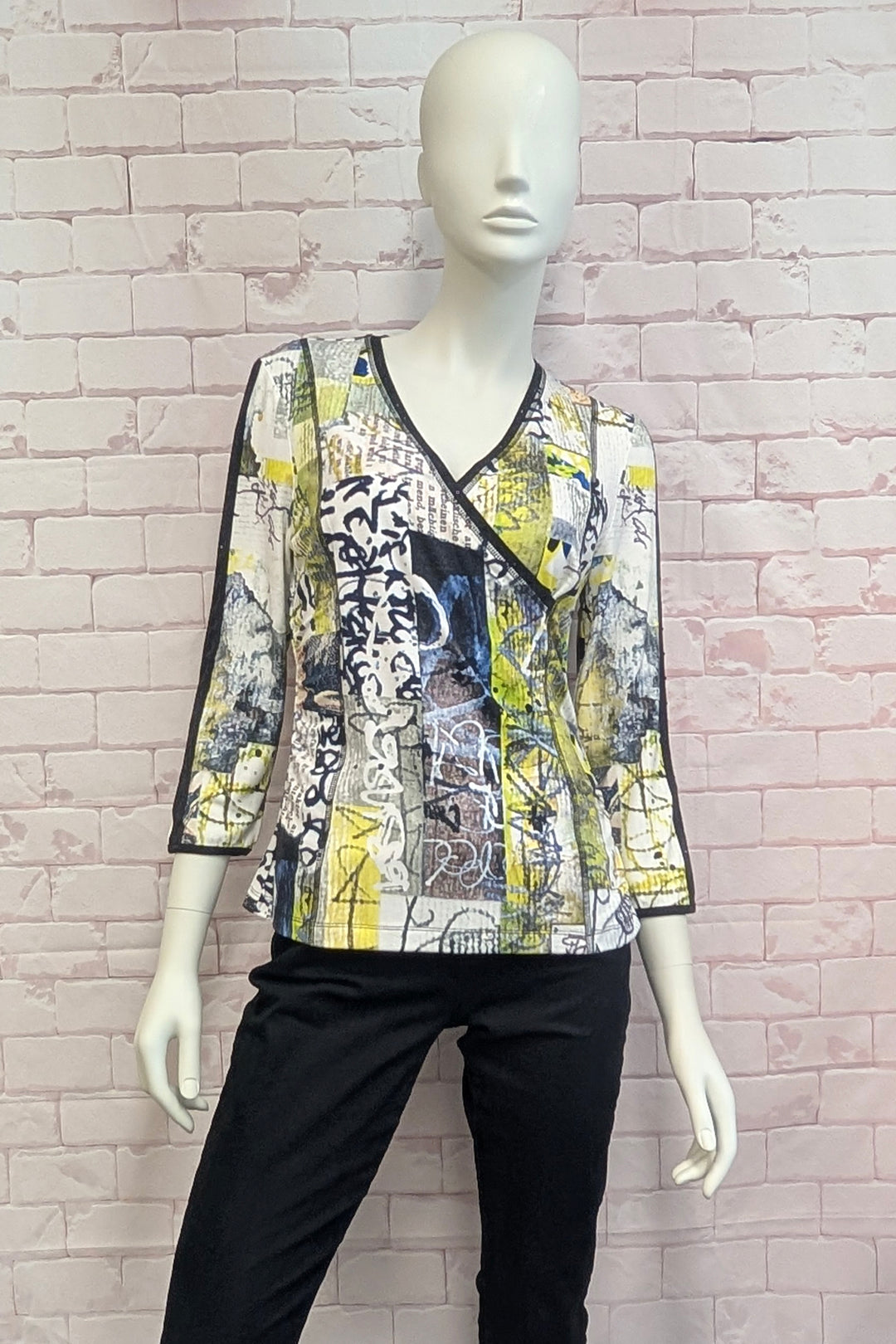 A women's wrap-style blouse with three-quarter sleeves, featuring a vibrant abstract collage print in yellow, black, white, and blue. The blouse is displayed on a white mannequin wearing black pants, set against a light pink brick wall background.