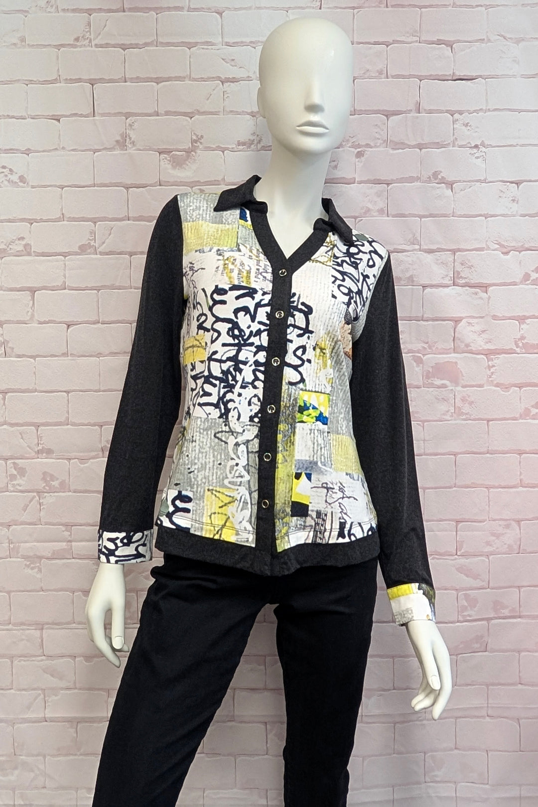 A long-sleeve button-front shirt with a black collar and cuffs, featuring a mixed abstract print panel in black, white, yellow, and gray, displayed on a white mannequin against a pink brick wall.