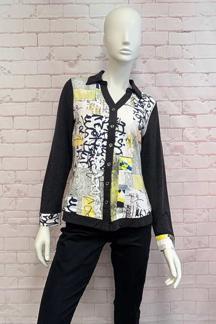 A long-sleeve button-front shirt with a black collar and cuffs, featuring a mixed abstract print panel in black, white, yellow, and gray, displayed on a white mannequin against a pink brick wall.