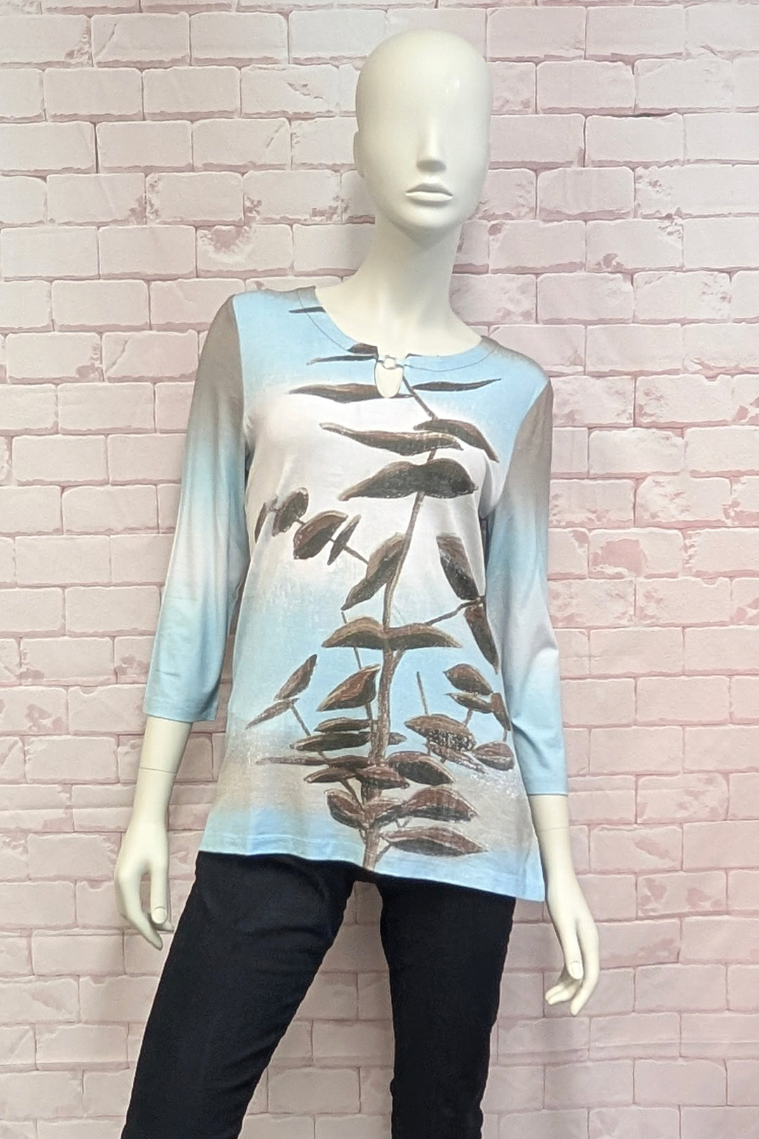 A  mannequin wearing a long-sleeve tunic top with a gradient design transitioning from light blue at the top to beige at the bottom, featuring a brown botanical leaf pattern printed down the front.