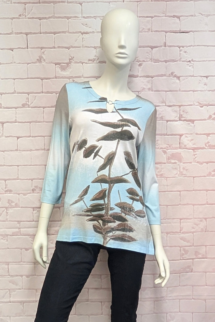 A  mannequin wearing a long-sleeve tunic top with a gradient design transitioning from light blue at the top to beige at the bottom, featuring a brown botanical leaf pattern printed down the front.