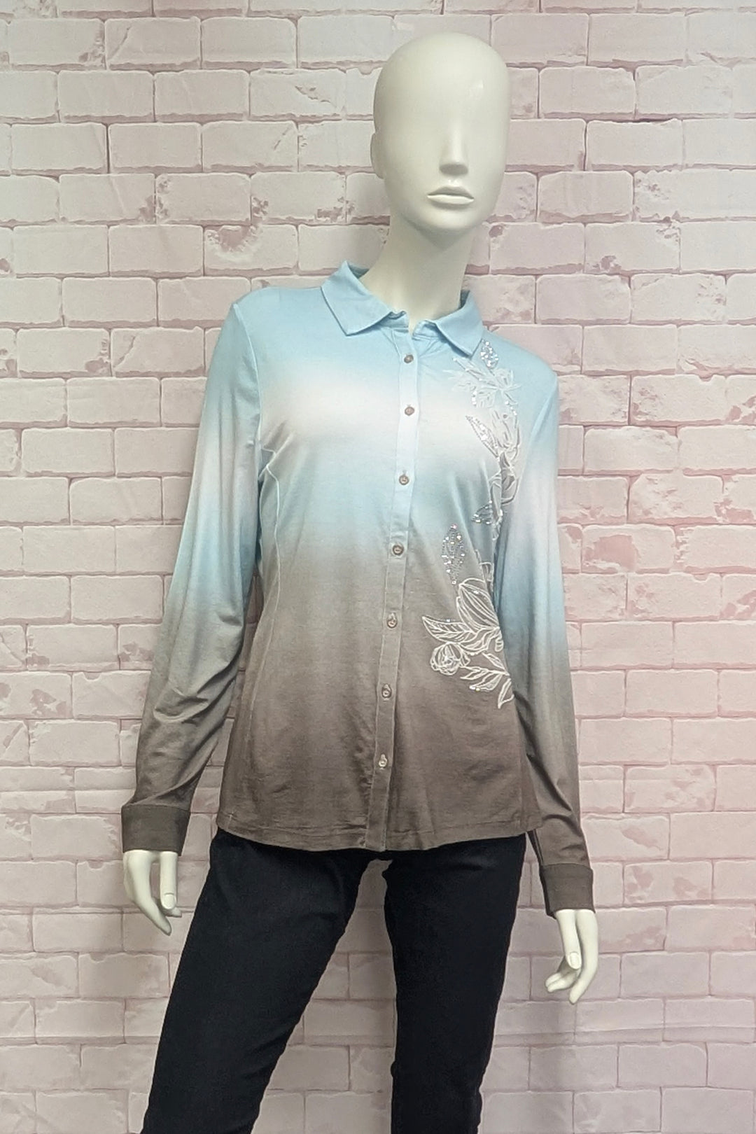 A mannequin wearing a blue and brown gradient blouse with flower embroidery shown on a brick background. 