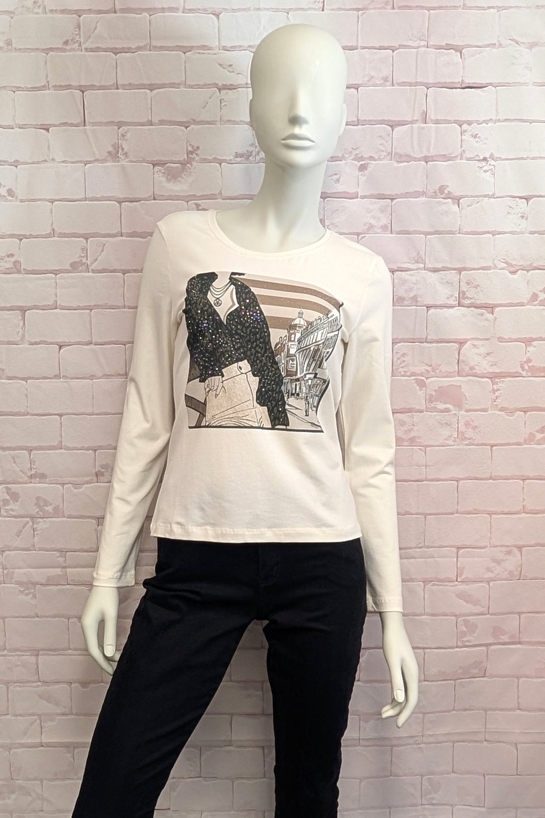 A mannequin wearing a cream long-sleeve graphic t-shirt with a black and white illustration of a woman, paired with black pants, set against a light pink brick wall background.