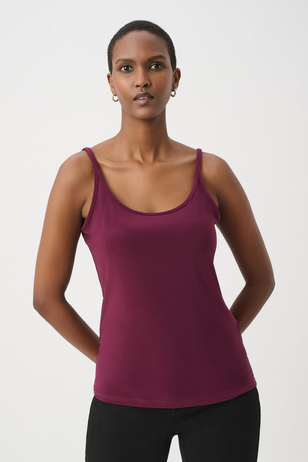 A woman modelling a purple coloured basic thin strapped camisole wearing black pants and shoes in front of a white background. 