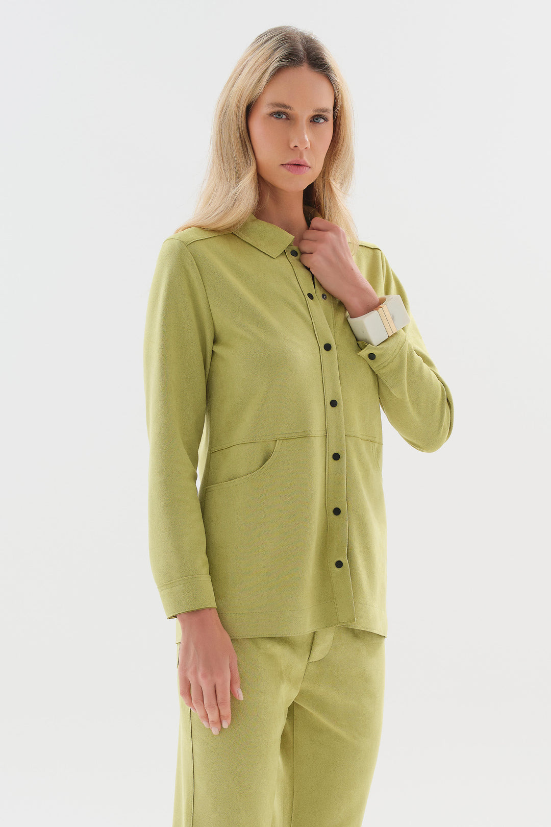 A woman modeling a matching light olive green two-piece lounge set, featuring a long-sleeve button-up shirt and coordinating trousers, against a plain light gray studio background.