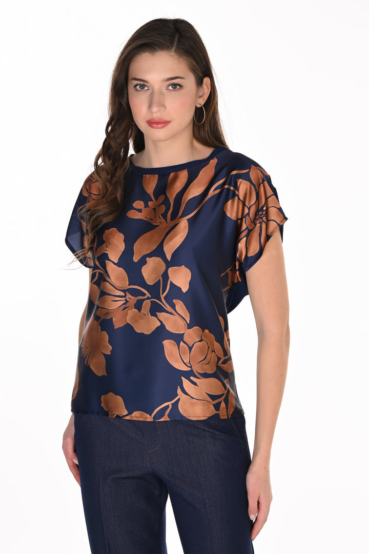 A woman modeling a navy blue short-sleeve blouse with a large brown floral print, paired with dark blue jeans, against a plain white background.