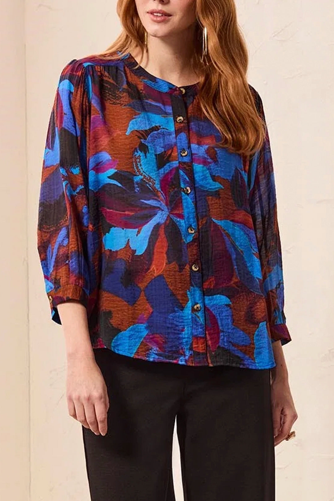 A woman wearing a long-sleeve button-front blouse with a bold abstract floral pattern in blue, purple, orange, and black, paired with black pants.