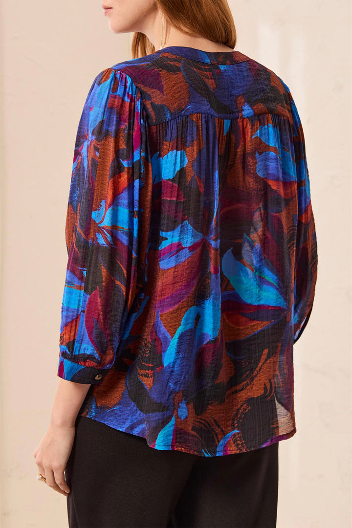 A woman shown from the back wearing a short-sleeve button-front blouse with a bold abstract floral print in blue, purple, brown, and black, paired with black pants against a light neutral wall.
