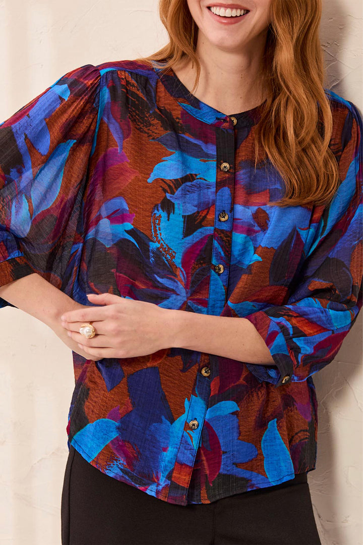 A woman wearing a short-sleeve button-front blouse with a bold abstract floral print in blue, purple, brown, and black, paired with black pants against a light neutral wall.