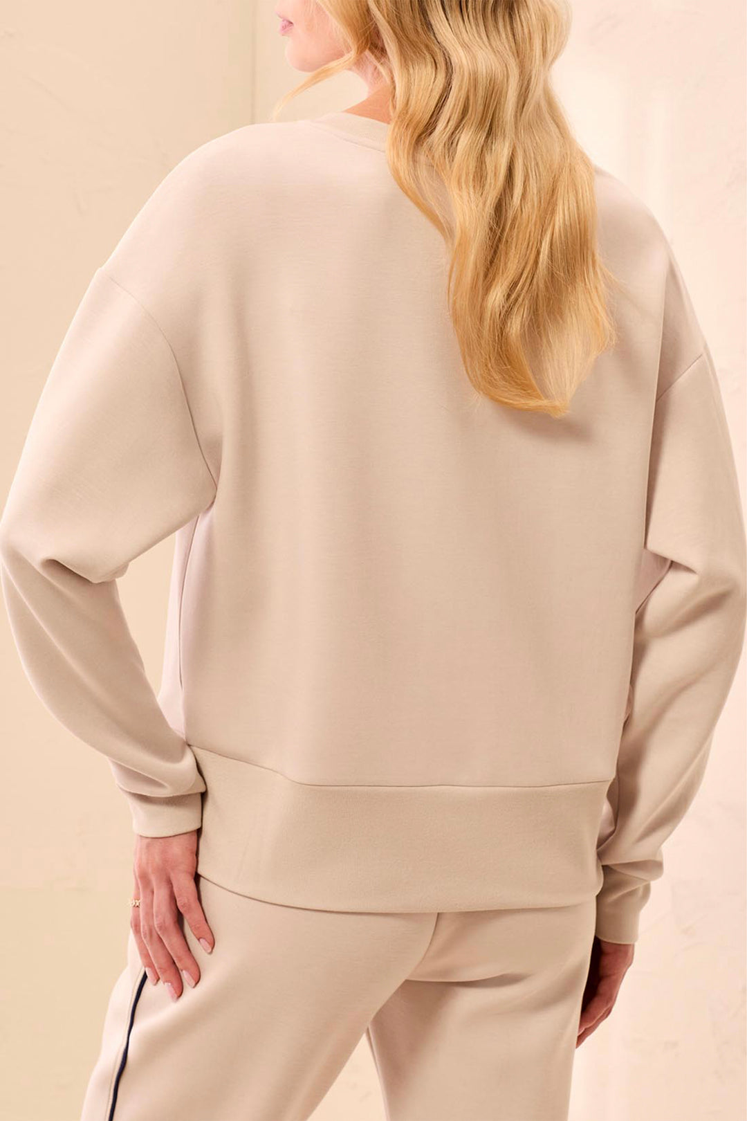 A woman shown from the back wearing a light beige crewneck sweatshirt and matching sweatpants against a soft beige studio background.