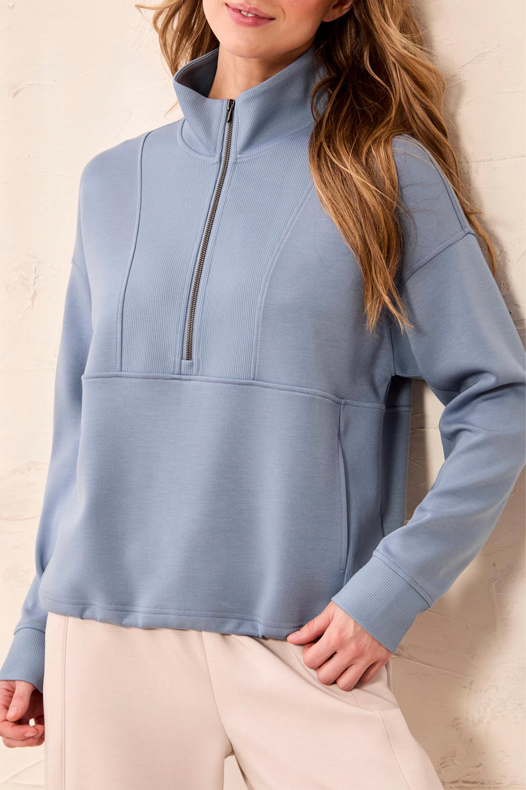 A woman wearing a dusty blue quarter-zip mock-neck sweatshirt and light beige pants, standing against a textured beige wall.