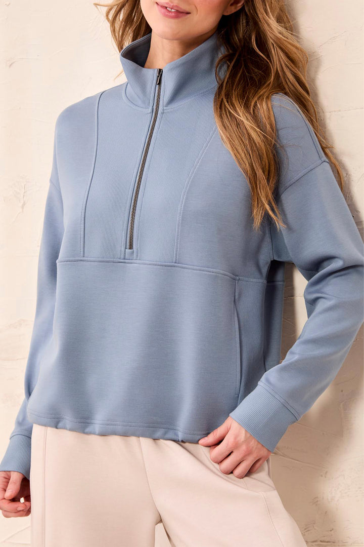A woman wearing a dusty blue quarter-zip mock-neck sweatshirt and light beige pants, standing against a textured beige wall.