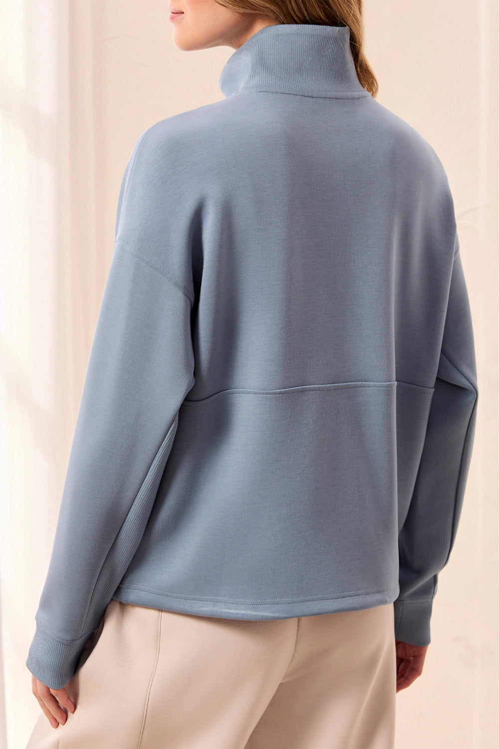 A woman shown from the back wearing a dusty blue mock-neck long-sleeve top and light beige pants, set against a soft, warm neutral background.