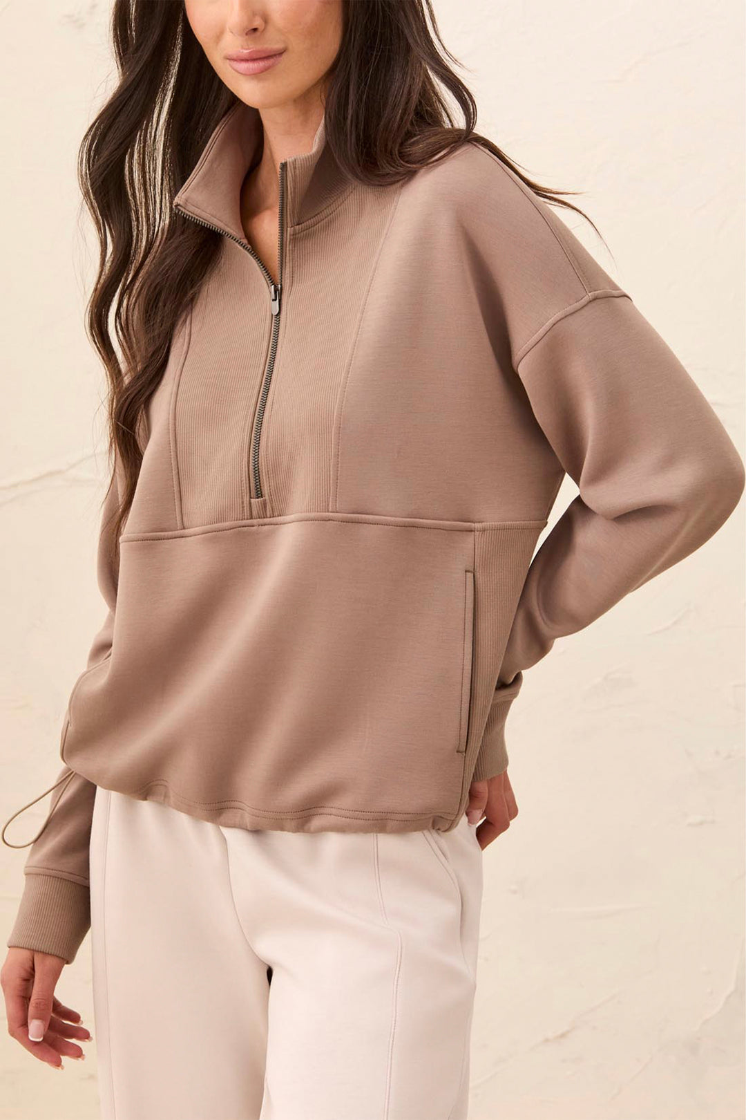A woman wearing a taupe brown quarter-zip pullover sweatshirt with a mock neck and zippered side pocket, paired with cream pants, against a textured beige wall.