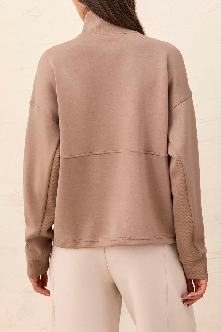 A woman shown from the back wearing a taupe-brown mock neck long-sleeve sweatshirt and light beige pants against a textured off-white background.