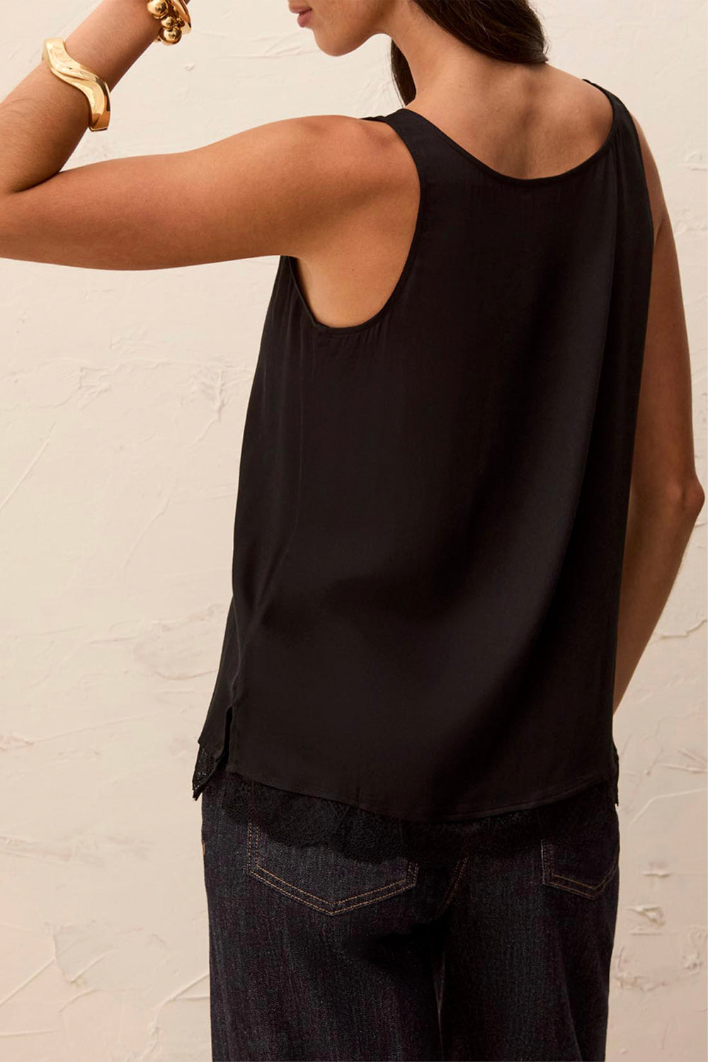 A woman wearing a black sleeveless top and dark jeans, shown from the back against a textured off-white wall.