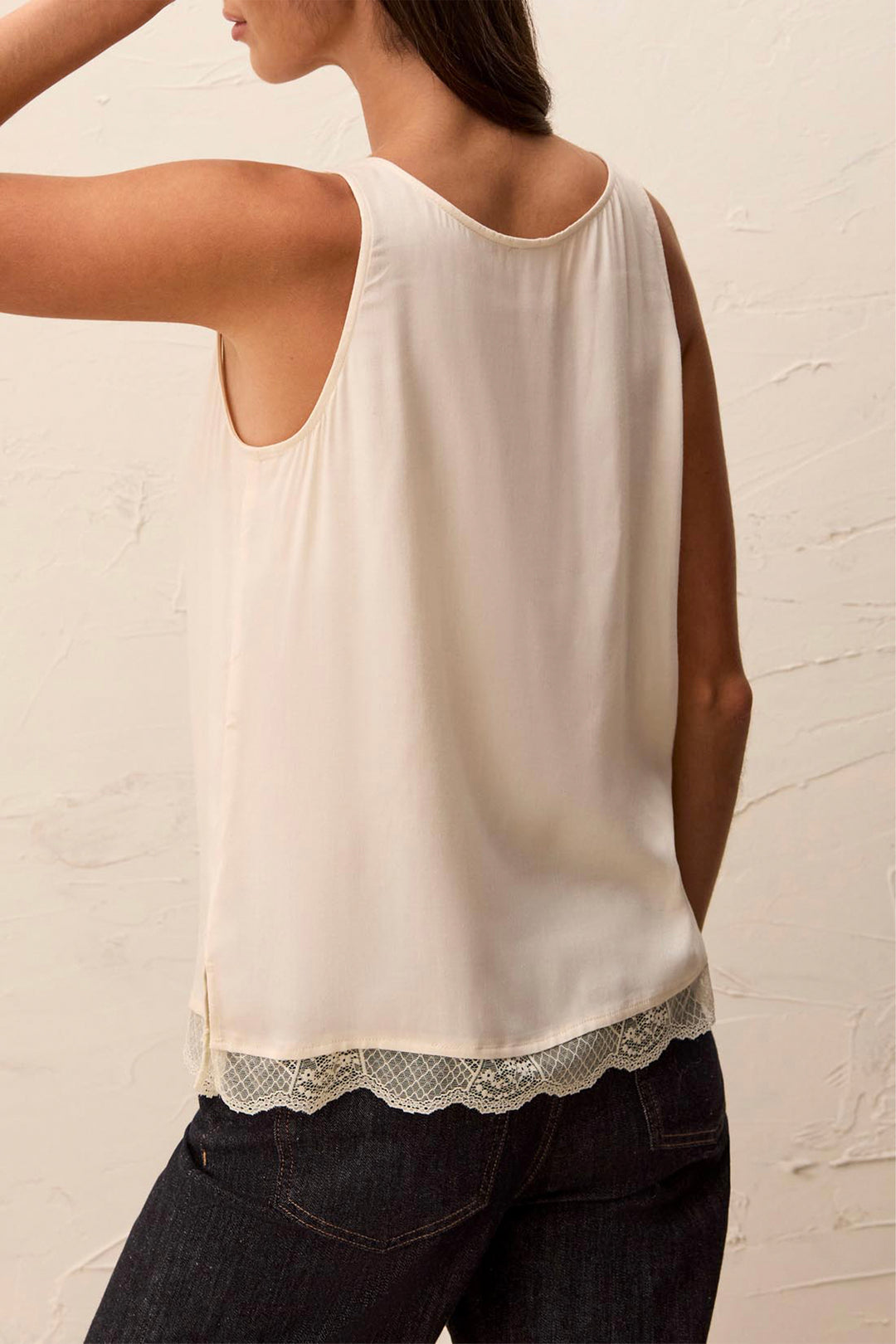 A woman wearing a sleeveless cream-colored top with a scalloped lace hem, paired with dark jeans, shown from a rear three-quarter view against a textured off-white wall.