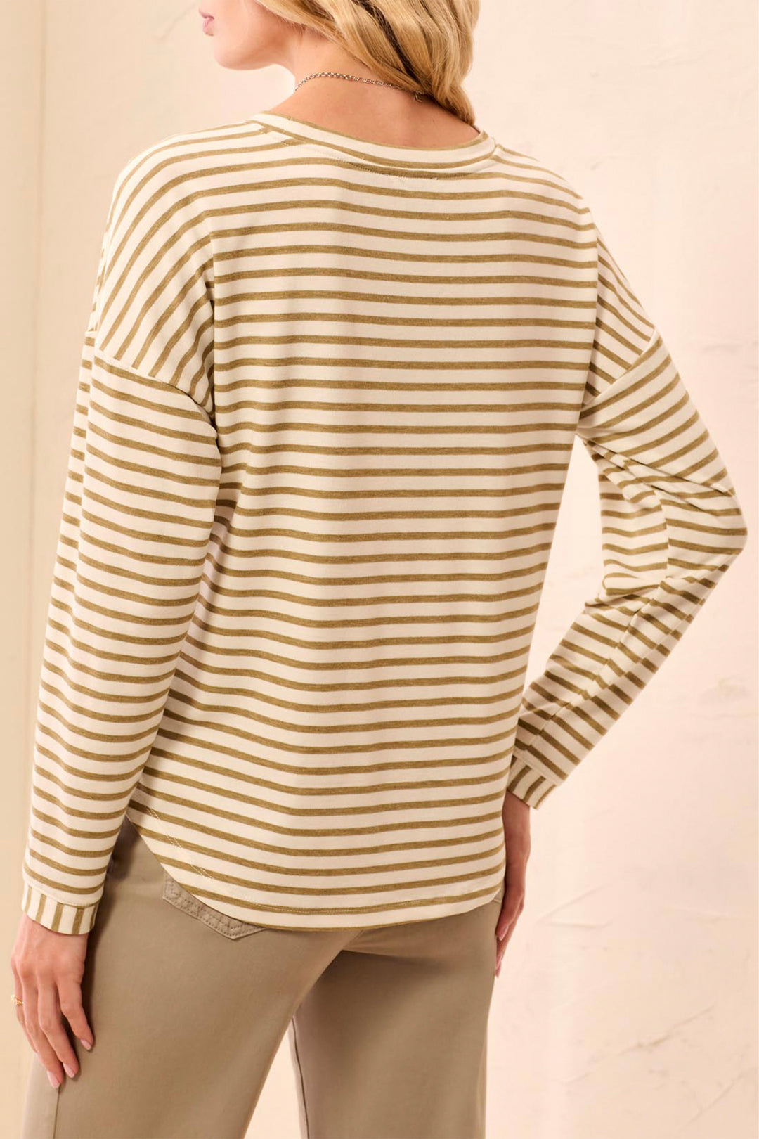 A woman shown from the back wearing a long-sleeve top with horizontal beige and white stripes, paired with khaki pants, against a light beige background.