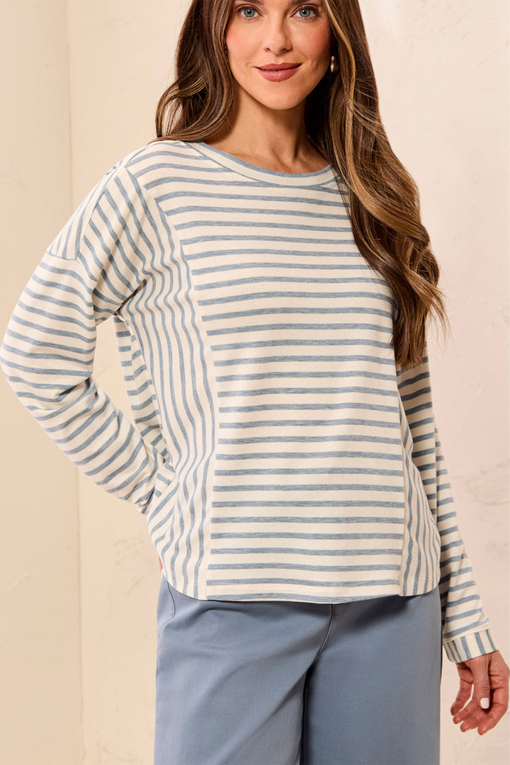 A woman wearing a long-sleeve striped top with horizontal stripes in light blue and off-white, paired with light blue pants, standing against a neutral beige background.