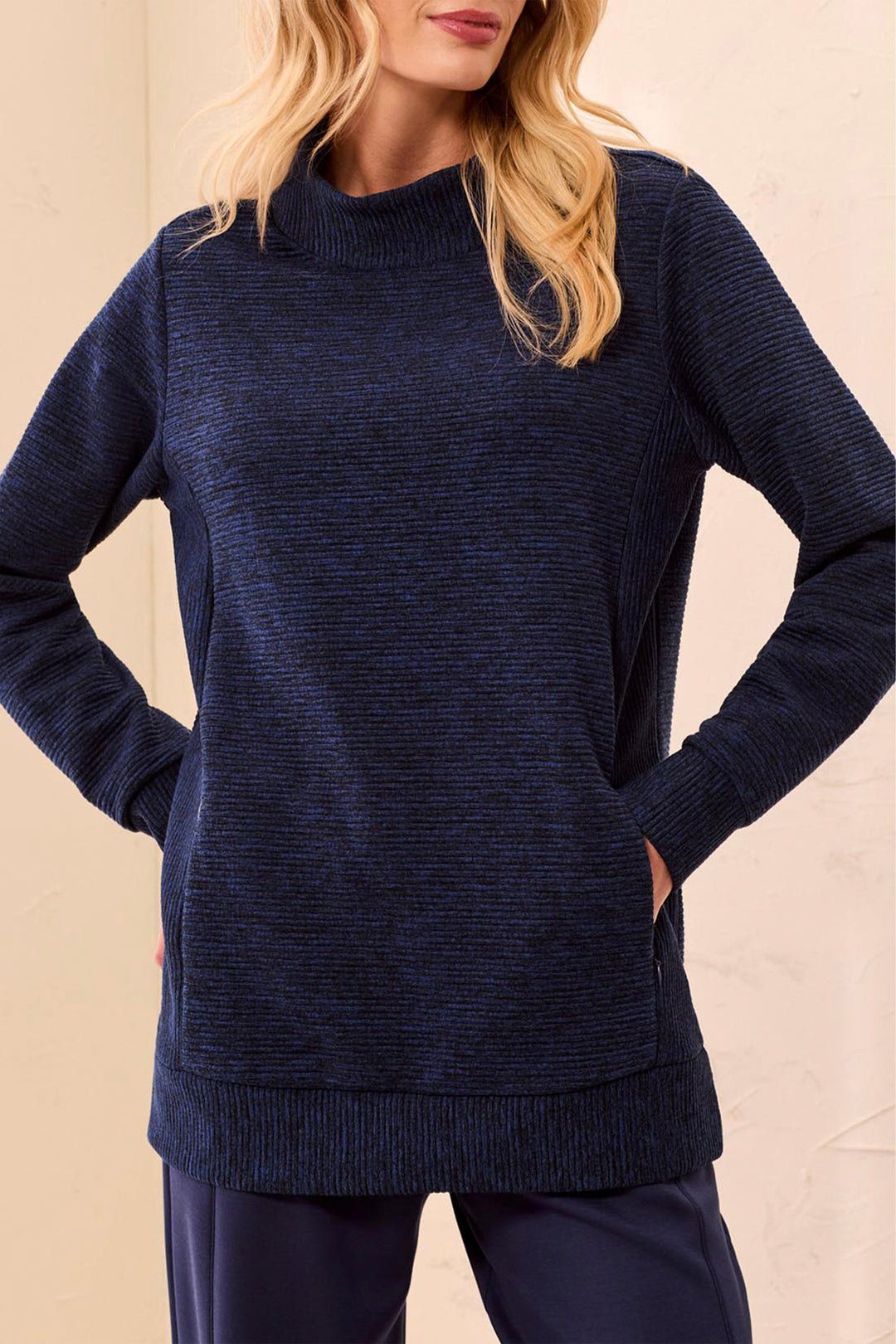 A woman wearing a dark navy blue ribbed knit mock neck sweater with side pockets, paired with navy pants, set against a warm beige background.