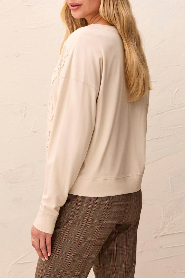 A woman shown from the back wearing a cream-colored long-sleeve crewneck sweatshirt and brown plaid trousers, standing against a textured off-white wall.