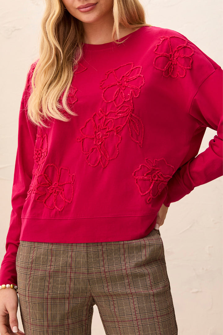 A woman wearing a bright pink crewneck sweatshirt with tonal floral embroidery and brown plaid trousers, photographed against a light textured wall.