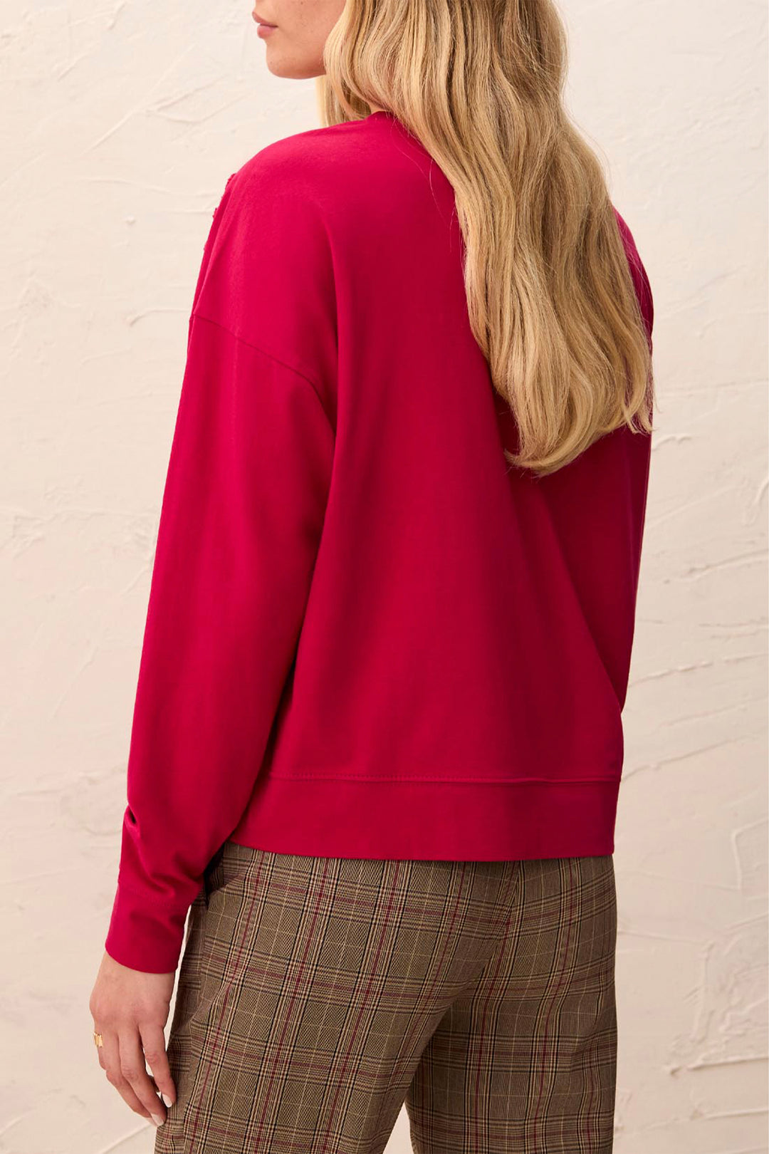 A woman shown from the back wearing a bright magenta crewneck sweatshirt and brown plaid trousers, standing against a textured off-white wall.