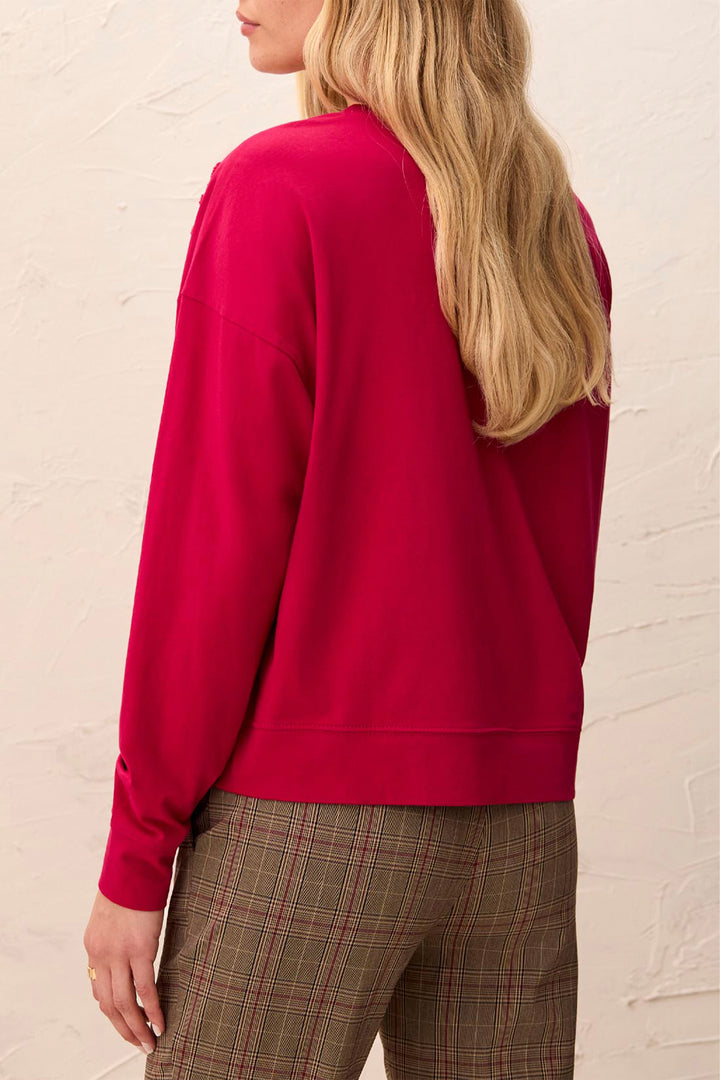 A woman shown from the back wearing a bright magenta crewneck sweatshirt and brown plaid trousers, standing against a textured off-white wall.