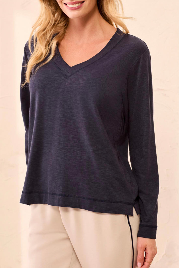 A woman wearing a dark navy long-sleeve top and blue jeans, standing against a textured beige wall.