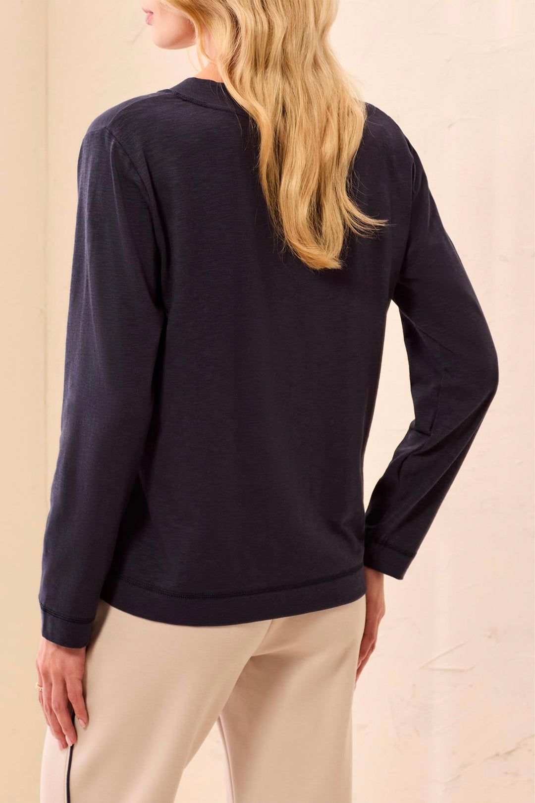 A woman shown from the back wearing a dark navy long-sleeve top and blue jeans, standing against a textured beige wall.