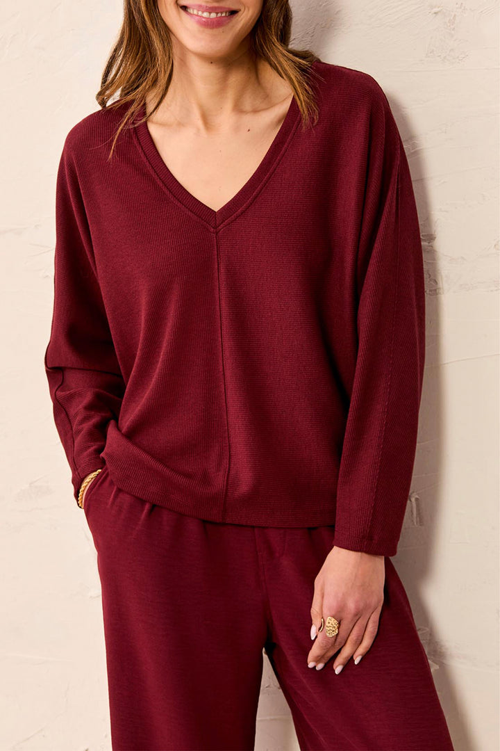 A woman wearing a matching burgundy knit lounge set consisting of a long-sleeve V-neck top and coordinating pants, standing against a light textured wall.