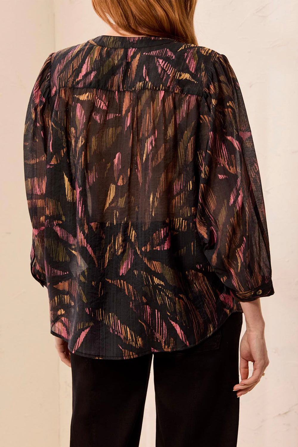 A woman shown from the back modelling a black leaf print loose blouse in front of a light beige background. 