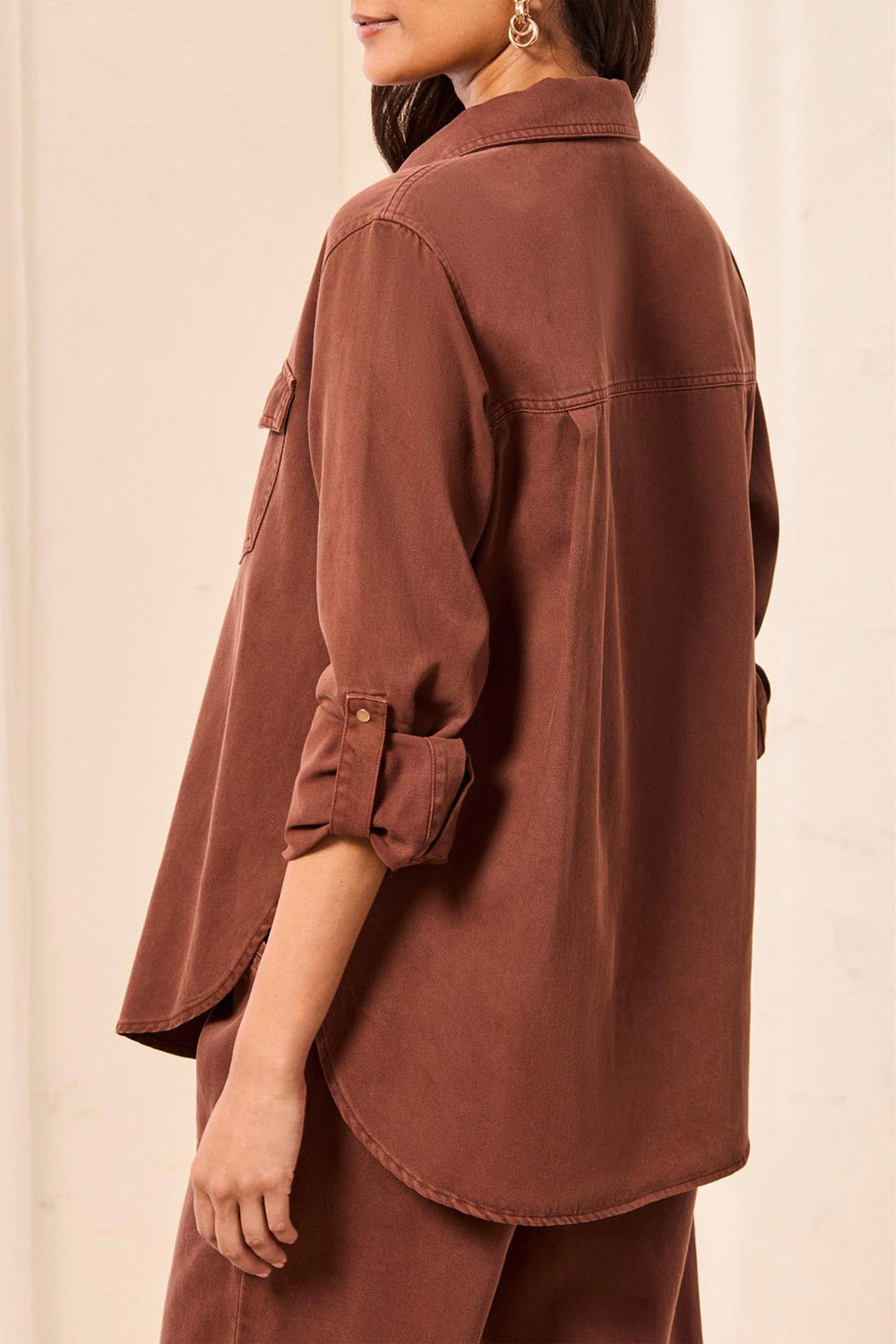 A woman shown from the back wearing a rust-brown button-up shirt with three-quarter sleeves and matching brown pants, shown against a plain light background.