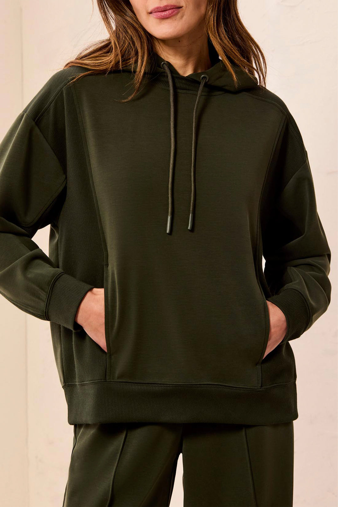 A woman wearing an oversized dark olive green hoodie with matching drawstrings and matching green pants, shown from the lips down against a plain light background.
