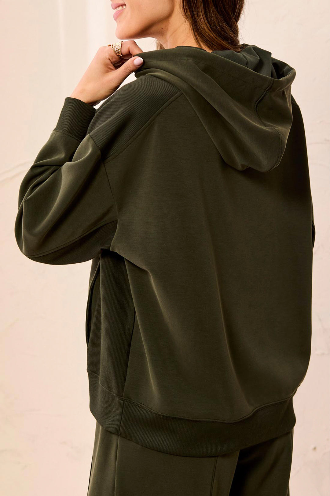 A woman wearing an olive green pullover hoodie, shown from the back and side, with her hand near the hood. The background is a clean, off-white textured wall.