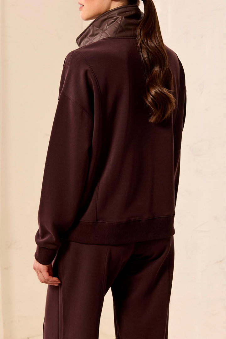 A woman shown from the back wearing a dark brown sweatshirt and matching sweatpants, with a quilted high-neck collar visible at the neck, against a plain light beige background.