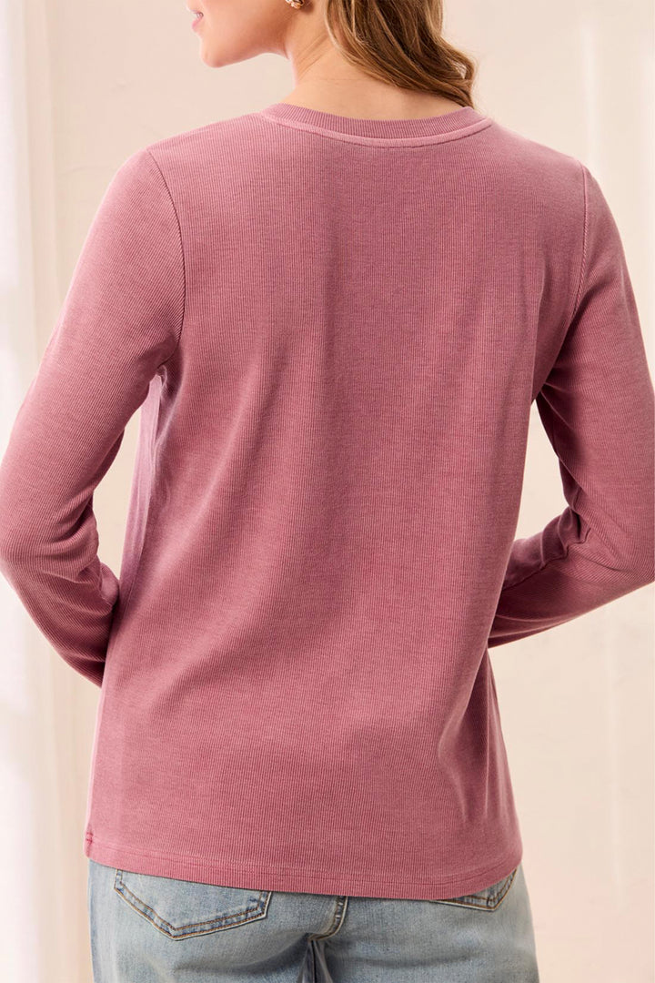 A woman shown from the back wearing a dusty rose long-sleeve crewneck shirt and light blue jeans, set against a soft beige indoor background.